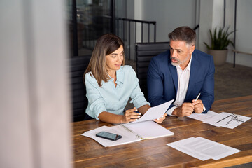 Diverse partners mature Latin businessman and European businesswomen discussing project with documents at table in office. Team of colleagues professional business people working together, copy space