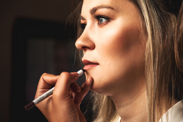 Make-up artist with lipstick pen doing makeup for lovely lady bride at wedding day in hotel room. Close-up of visagiste make wedding makeup for woman. Concept of luxury wed service. Copy ad text space