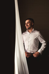 Confident guy groom in white shirt in hotel room posing looking at window. Portrait of young man...