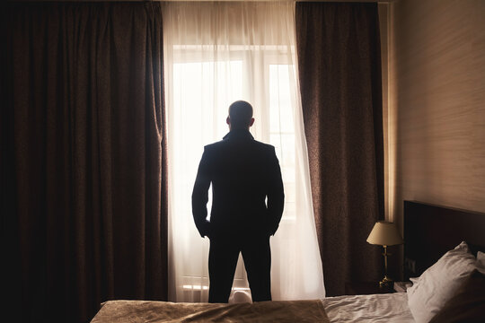 Silhouette of confident guy businessman in black suit standing at window in hotel room, rear view. From behind man getting ready to meet his bride. Concept of business married. Copy ad text space