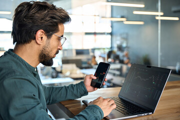 Stock trader investor broker holding mobile phone looking at laptop using computer analyzing trade cryptocurrency financial digital market indexes crypto stockmarket charts data, side view. - obrazy, fototapety, plakaty