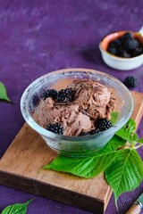 Two scoops of chocolate ice cream in a glass bowl, topped with fresh blackberries and chocolate chips, on a purple concrete background. Homemade ice cream recipes.