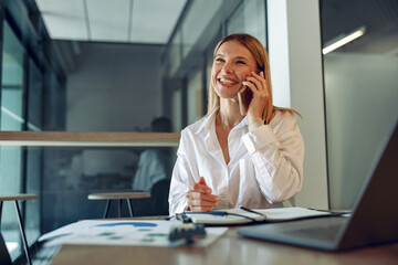 Woman sales manager talking by phone with client sitting in modern coworking and working on laptop