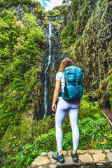 Naklejka na ściany i meble Sporty woman with backpack standing on stone wall viewpoint and watches green overgrown Risco waterfall. 25 Fontes Waterfalls, Madeira Island, Portugal, Europe.