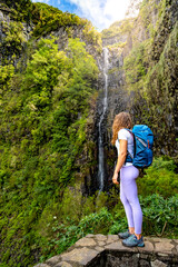 Naklejka na ściany i meble Female tourist with backpack standing on stone wall viewpoint and watches green overgrown Risco waterfall. 25 Fontes Waterfalls, Madeira Island, Portugal, Europe.