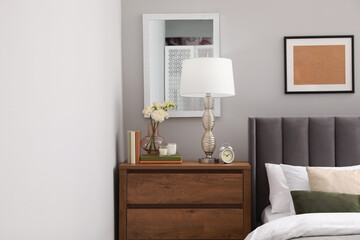 Comfortable bed with cushions, lamp and different decor on wooden chest of drawers in room. Stylish interior