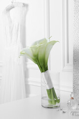 Beautiful calla lily flowers tied with ribbon in glass vase, bottle of perfume and jewelry on white table