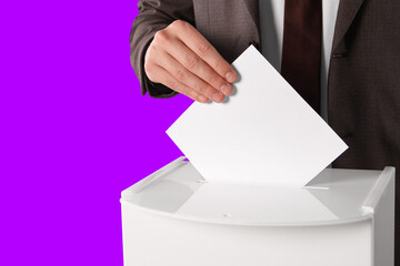 Man putting his vote into ballot box on violet background, closeup