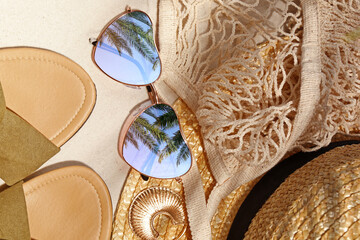 Flat lay composition with stylish sunglasses and other fashionable accessories on sand. Sky and palm trees reflecting in lenses