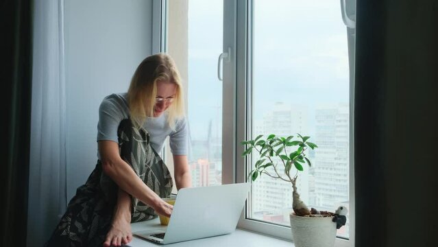A happy barefoot woman using a laptop while sitting on a windowsill in a cozy apartment with a city view. Remote work, e-learning, freelance.