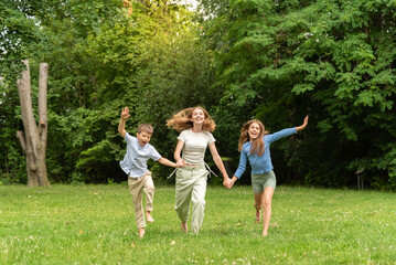 Naklejka na ściany i meble Three happy children running holding hands in forest outdoors. Two happy blonde teens and a little boy rejoice and run in the meadow. Happy childhood family and friendship concept. Children, lifestyle