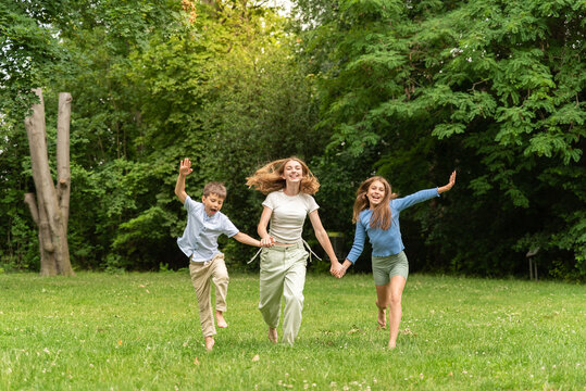 Three happy children running holding hands in forest outdoors. Two happy blonde teens and a little boy rejoice and run in the meadow. Happy childhood family and friendship concept. Children, lifestyle
