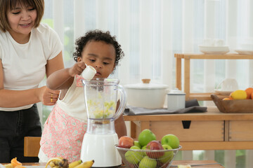 Little mixed race child girl, African and Asian, and her mother making healthy fruits smoothie together in kitchen at home. Cute daughter helping mom pouring fresh milk into blender