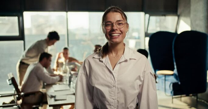Portrait of a happy girl in glasses and a white shirt in the office, who smiles and looks at the camera. Posing against the backdrop of the working process of office workers