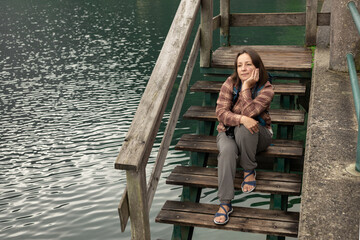 Young female photographer with backpack sitting on wooden descent to the water and admiring the...