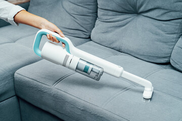 A woman is vacuuming a sofa. Manual vacuum cleaner for furniture cleaning. House cleaning. Stylish white vacuum cleaner.
