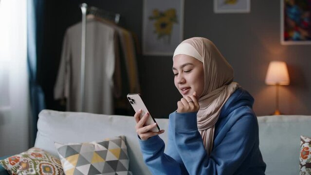 Smiling modern young muslim woman in hijab scrolls social networks on smartphone. Smiling young modest female looking at digital device while browsing social media online at modern positive apartment