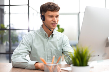 Cheerful young support phone male operator in headset, at workplace while using computer, help...