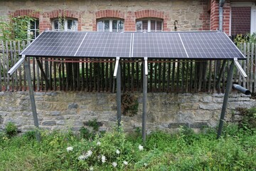 Solaranlage über alter Backsteinmauer, Holzzaun und Blumenbeet vor altem rot-braunem backsteinhaus mit Rundbogenfenstern auf Bauernhof am Nachmittag im Sommer 