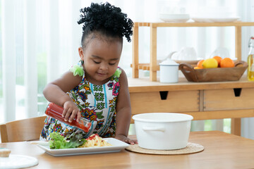 Little African toddler girl pouring ketchup on spaghetti with vegetables in kitchen at home. Happy...