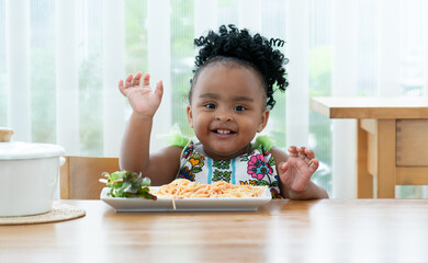 Portrait of little African toddler girl smiling while eating spaghetti and vegetables with hand in...