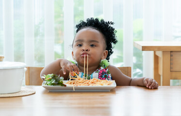 Portrait of little African toddler girl eating spaghetti tomato sauce and vegetables with hand in kitchen at home. Happy messy preschool child eating fresh cooked healthy meal with yummy face