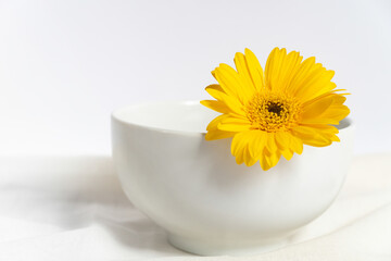 Eine weiße Porzellanschüssel aus der eine gelbe Blüte herausguckt auf einem weißen Tuch,...