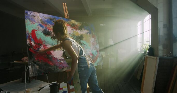 Millennial girl artist paints picture in loft studio puts paint on canvas with her hands. Abstract cinematic. Women's power, freedom of expression, Capacity Development, Lifestyle, light rays