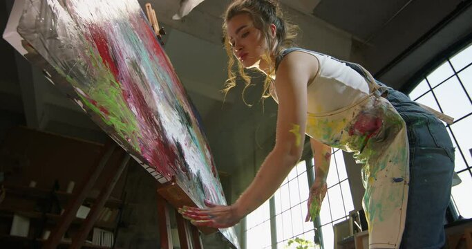 Millennial girl artist paints picture in loft studio using her hands to apply paint to canvas. Abstraction cinematic. Leaning over a bucket of paint to pick up paint in their hand, non-standard view