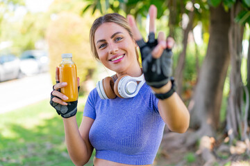 Young pretty sport woman holding an orange juice at outdoors smiling and showing victory sign