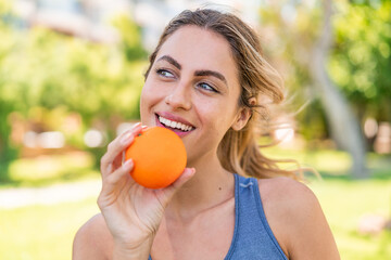 Young blonde woman at outdoors holding an orange with happy expression