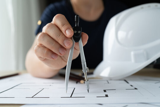 Female architect, interior designer holding compass drawing tool. Woman working on home floor plans, building blueprint projects. Hard hat safety helmet lying on the table. Design studio desk scene.