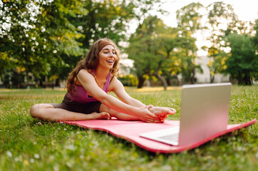 Beautiful woman practices yoga with a laptop via video link in the morning or in the park at sunset. Sportswoman is engaged in stretching on a gymnastic carpet on a green lawn. Fitness.