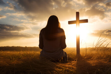 Silhouette of a woman sitting on the grass praying in front of a cross at sunset - Powered by Adobe