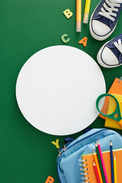 Embracing the journey of knowledge. Top view vertical photo of assorted colorful school materials, pair of shoes, rucksack, colorful letters on green background with blank circle for promo or text