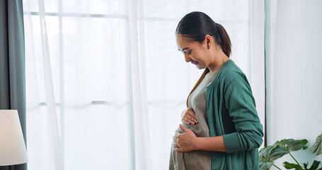 Happy Asian pregnant woman lovingly touching her unborn child while standing by the window of the house.