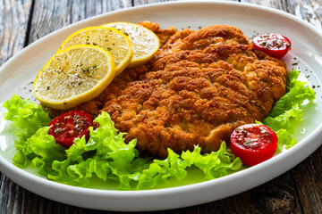 Crispy breaded seared chicken cutlet with lemon slices and fresh vegetables on wooden table
