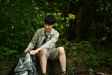 Man with backpack dressed in active trekking clothes sitting on dried tree trunk while exploring nature.