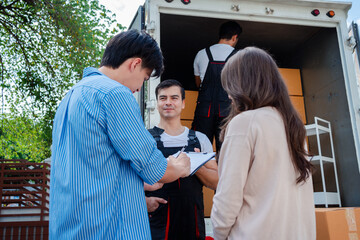 Obraz premium Asian Couple check while unloading boxes and furniture from a pickup truck to a new house with service cargo two men movers worker in uniform lifting boxes. concept of Home moving and delivery.