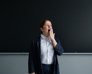 Caucasian female teacher in a suit yawns at the blackboard.