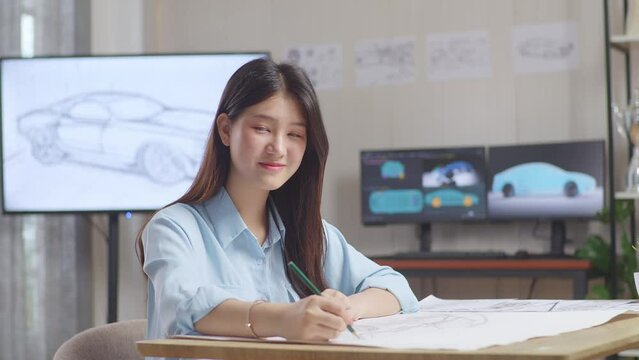 Asian Female Smiling And Showing Thumbs Up Gesture To The Camera While Working On A Car Design Sketch On Table In The Studio With Tv And Computers Display 3D Electric Car Model 
