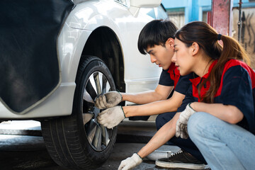 Selective focus of side view of two young Asian mechanics, man and woman, in uniforms and gloves, squatting to install or remove or replacing a lug nut of an automobile front wheel by hand in a garage