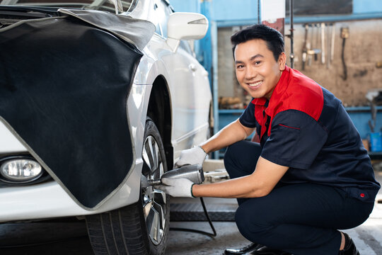 Selective focus of a mid-adult Asian male mechanic in uniform and gloves, squatting using a pneumatic wrench changing lug nuts of a car front wheel while turning to smile at the camera in the garage.