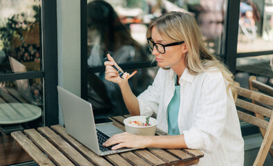 A woman eats a salad and works on a laptop in a cafe.