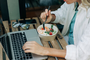 Unrecognizable woman eats salad and works on a laptop in a cafe.
