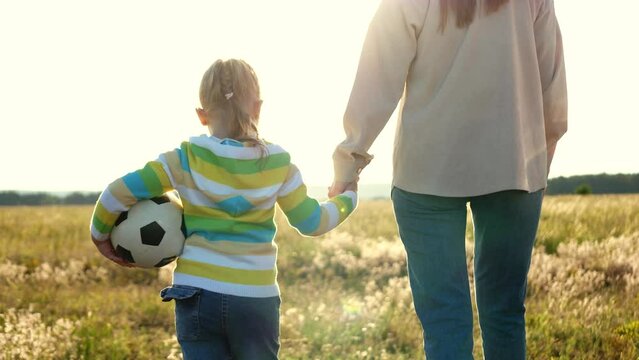 mother holds girl daughter by hand walk. girl walk with soccer ball. child siotrit smiling mother. happy family. chidhood dream. walk children football field sunset. family happiness concept. children