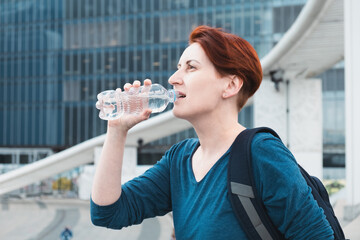 Middle-aged woman with a short haircut drinks water from a plastic bottle against the backdrop of a modern urban business district. The need to drink fluids while in the heat.