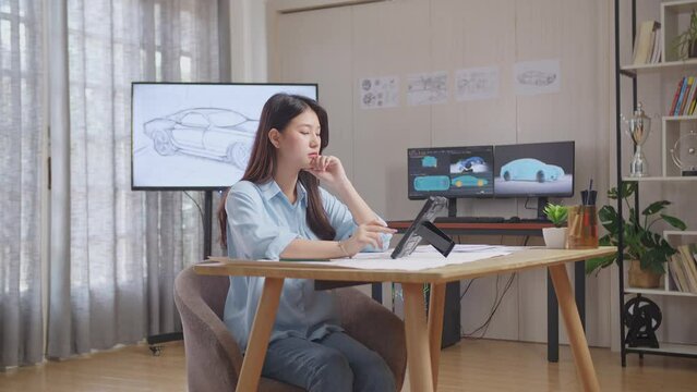 Side View Of Asian Female Thinking About New Car Concept While Working On A Car Design Sketch On Tablet In The Studio With Tv And Computers Display 3D Electric Car Model 
