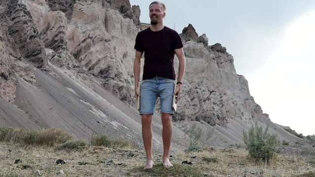An adult young slender guy in shorts and a T-shirt prepared to meditate on a Sadhu board in the picturesque mountains on a very windy day. Cheerful thin long-haired man likes to meditate outdoors