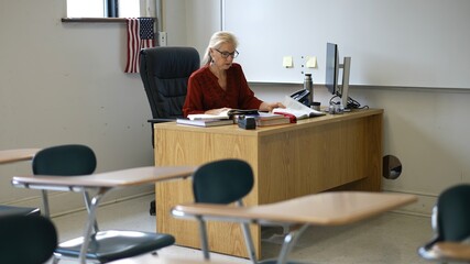 Happy woman teacher sitting at desk in empty school classroom desk grading papers working on assignments. US American flag in the background.